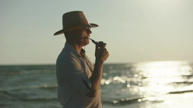Man wearing sunglasses and straw hat standing in windy weather on beach and puffing pipe. Summer at seaside.