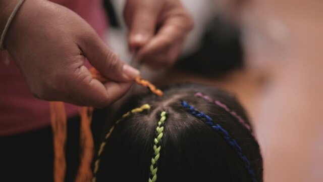 Close-up VDO: Cute long, black hair Asian girl is being braided in multi-colored threads by woman's hand in beauty salon. Child-like fashion, weaving pigtails with decoration yarn or knitting wool.