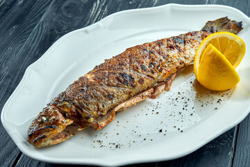 Whole grilled trout on charcoal fire, served in a white plate with lemon on a dark wood background.