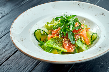 Appetizing, dietary salad with lettuce, cucumber, avocado and lightly salted salmon in a white plate on a dark wood background. Healthy food