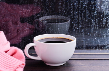 A hot white coffee cup and pink sweater on a vintage wooden window sill or a window table with raindrops on a glass background.