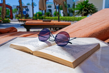 A close view of an open book and sunglasses on the sun loungers by the pool. Behind there are sun umbrellas, resort buildings, garden. Vacation concept, reading outdoors, recreation by the sea.