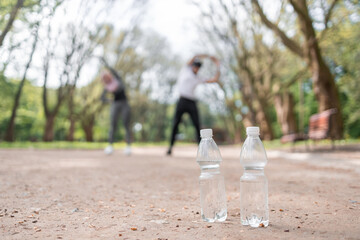 Obraz premium Plastic bottles with fresh cold water standing on road with blur background of muslim couple stretching body. Young man and women training together outdoors. Refreshing concept.