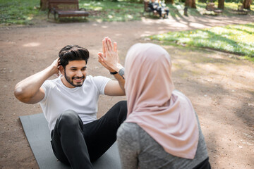 Obraz premium Muscular hindu guy training abdominal muscles on yoga mat with help of woman in hijab that holding his feet. Active young couple training together on fresh air.