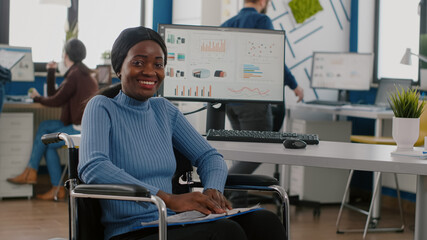 Happy african businesswoman with disabilities looking at camera smiling in immobilized paralysed in wheelchair in business economic office, working on pc with data prosessing financial information