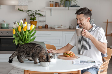 Young man with cute cat drinking coffee in kitchen