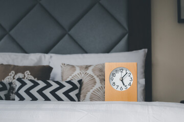 A wooden framed alarm clock rests on a white bed adorned with lovely pillows behind it.