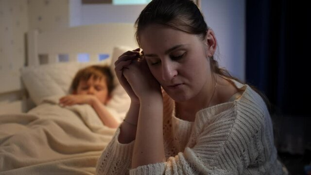 Upset and stressed mother crying next to bed of her little son sleeping at night. Concept of lonely mother, family problems and maternal depression
