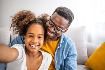Fototapeta premium Family time. Happy afro father taking selfie with his cute daughter at home, sitting on sofa. Happy african american man and his daughter taking selfie. Family time.