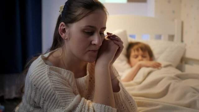Young mother sitting next to bed of her sleeping son suffering from depression and bad thoughts. Concept of lonely mother, family problems and maternal depression