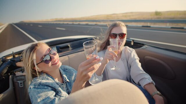 Two young woman ride in back seat of convertible drink champagne from glasses and enjoy life, against background of highway. Young women in sunglasses drink champagne on driving car without roof