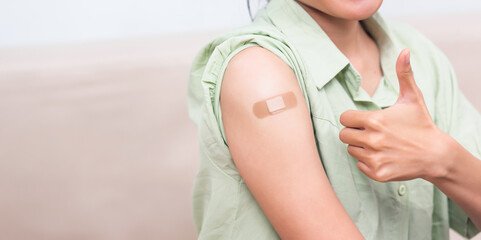 Asian young woman showing gesturing thumb up or like and her arm with plaster band aid after coronavirus Covid-19 vaccine injection on shoulder, coronavirus COVID-19 vaccination concept - obrazy, fototapety, plakaty