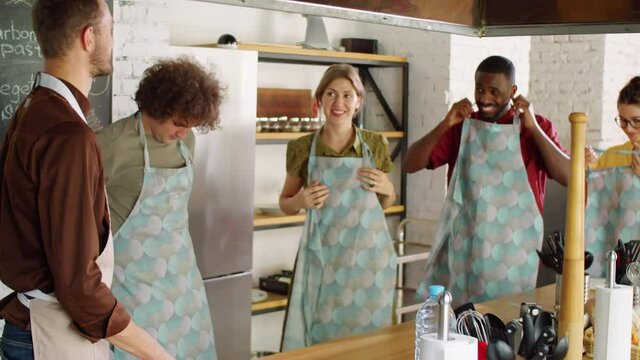 Group of cheerful multiethnic men and women smiling and chatting with chef while putting on aprons before culinary master class