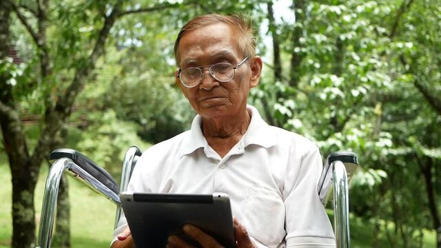 Senior man in wheelchair using digital tablet, looking at screen, reading e-book, chatting online with grandchild, watching movie in the park.