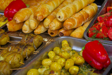 Various prepared foods are on the trays. Potato slices on skewers, sausages in dough, red bell peppers and other products. Food cooked over an open fire offered at a street food fair, festival.