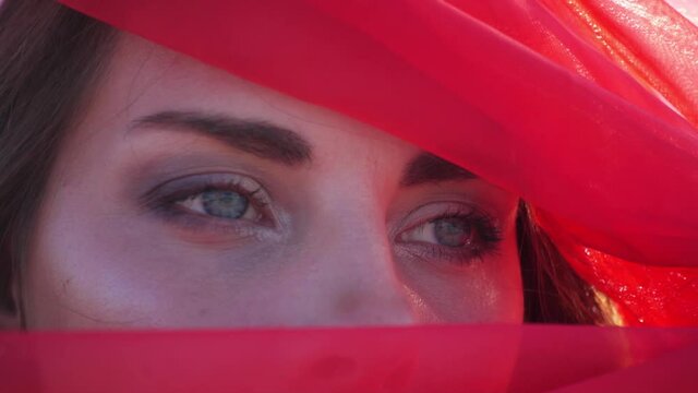 Cinematic close-up portrait face blue eyes young woman girl model in red scarf burqa hides her face. Beauty, fashion abstract film. Concubine. Red sun of desert. Shaman mystical man. Accept fate