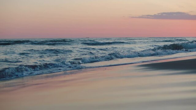 Low angle view of sea waves rolling and splashing on sandy beach. Pink twilight sky. Summer evening at seaside.