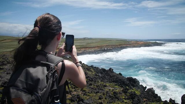 Woman taking a photo with her mobile phone at coastline in the Big Island of Hawaii. Mid angle, parallax movement, slow motion, HD.