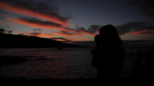 Woman photographing sunset clouds at pink hour in Keanae Peninsula, Maui, Hawaii. Mid angle, parallax movement, slow motion, HD.