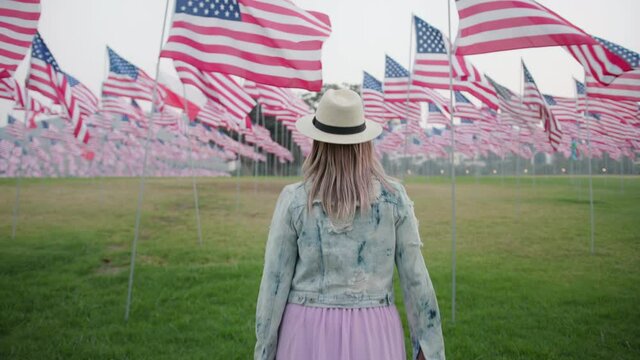 Camera following young stylish woman in memorial park with many US flags flattering by the wind on the poles. Model walking by the green meadow between National American flags with stripes and stars