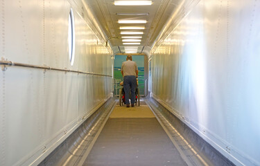 View through door into empty narrow airbridge, man walking with woman in wheelchair