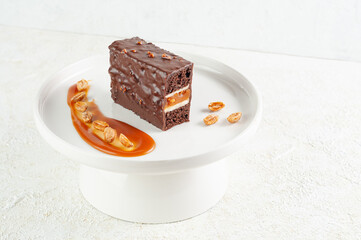 Snickers cake. Dessert based on chocolate biscuit, creamy mousse with caramel. Decorated with caramelized peanuts. On a white plate. Light background.