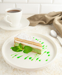 Pistachio cake. With airy sponge cake and custard and pistachio cream. Decorated with topping and a sprig of mint. On a white plate. In the background there is a napkin and a cup of tea.