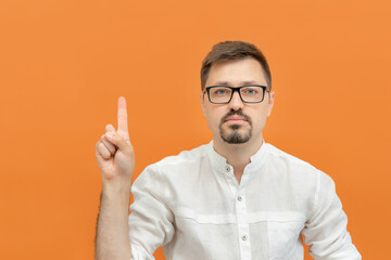 Young bearded thoughtful man in eyeglasses pointing his finger up indicates to copy space, orange studio background