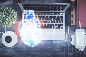 Heart hologram drawings over computer on the desktop background. Top view. Multi exposure. Concept of medical education.