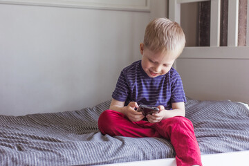 A blond boy sits on the bed and holds smartphone in his hands. Online communication, video communication, education