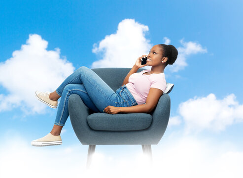 Comfort, People And Furniture Concept - Happy Smiling Young African American Woman Calling On Smartphone Sitting In Modern Armchair Over Blue Sky And Clouds Background