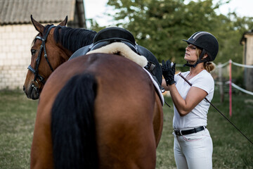 Obraz premium Иautiful woman dressed in sports clothes is preparing for training session on horse. Summer evening.Сoncept of active recreation. .