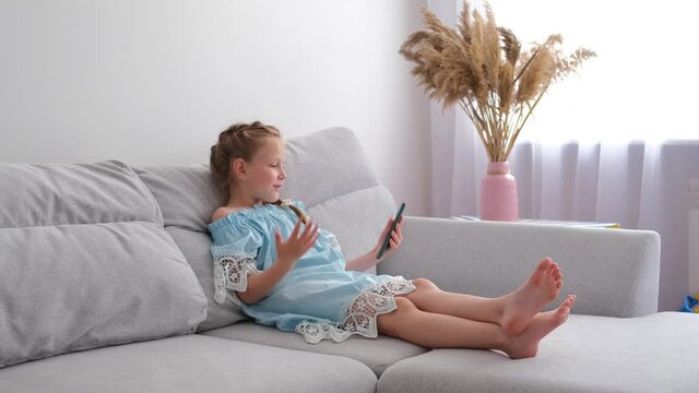 A little girl is sitting on the couch and talking on the phone. Online communication via video communication.