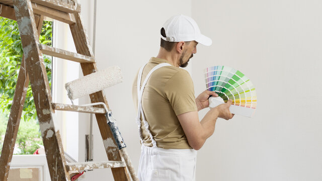 house painter man decorator choose the color using the sample swatch, work the white wall of the house to renovate, a wooden ladder, a paint roller and a green window as a background