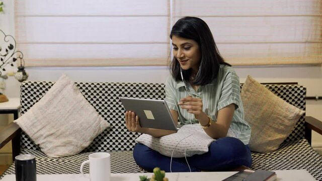 Beautiful Indian girl holding digital tablet watching funny videos,movie or social media content while sitting in home office.Pretty woman looking at tablet screen and watching something funny indoor.