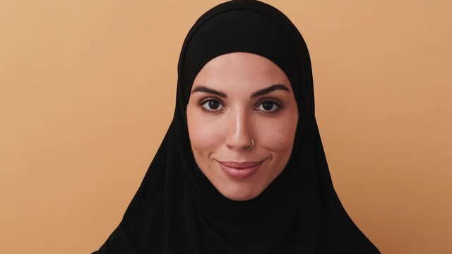 A close-up view of a beautiful arabian woman wearing a black hijab is looking to the camera standing isolated over a beige wall in the studio