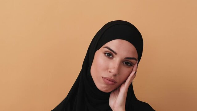 A close-up view of a beautiful arabian woman wearing a black hijab is thinking about something standing isolated over a beige wall in the studio
