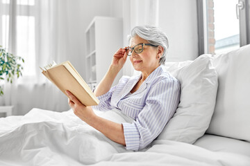old age, leisure and people concept - happy smiling senior woman in glasses reading book in bed at...