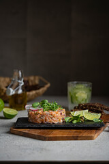 Salmon fish tartare with micro greens, lime on black stone plate, dark background. Grey textured table, healthy sea food concept. Macro shot with copy space.