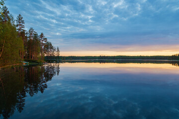 Fototapeta na wymiar Beautiful view of quiet forested shoreline, cloudy sky and their reflections on a calm lake at sunset in Finland at summer.