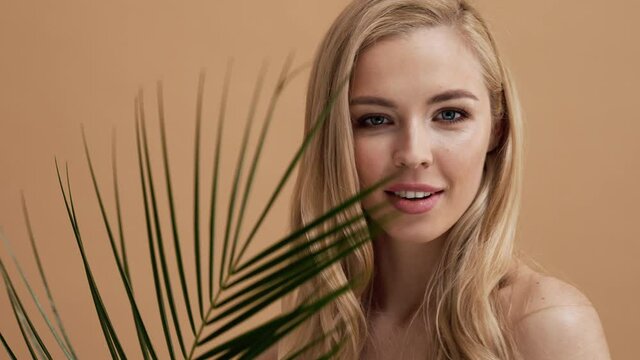 A beautiful blonde woman is posing with palm leaves standing isolated over a beige wall in the studio