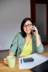 Stylish brunette woman in glasses sitting at wooden table with notepad and having phone call