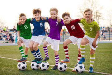 Group of happy junior soccer boys in colorful jersey uniforms. Five joyful kids from different countries with soccer football balls. Children in opposite teams huddling together on a grass pitch