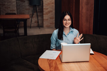 Woman sitting cafe coffee break open space work laptop have lunch documents papers. Lady dressed striped shirt stylish glasses speaking with colleagues video voice chat waving smiling copyspace