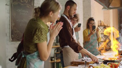 Group of excited multiethnic students taking pictures with smartphones as chef flambeing food in pan during cooking master class