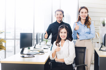 Asian customer service team looking at the camera and doing thumb up in call center. Group of young smiling operator with headset in modern office. Copy space