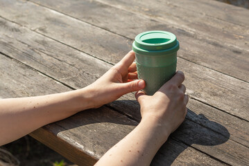 A girl holds green reuse cup in her hands against the background of a wooden table, sunlight, copy space