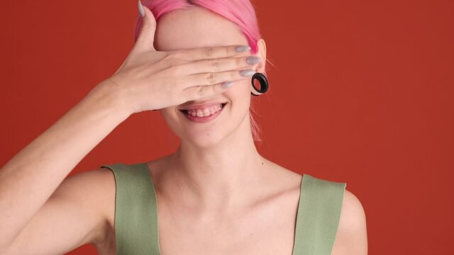 A beautiful young woman with pink hair is showing buddhism gestures of three monkeys standing isolated over orange wall in the studio