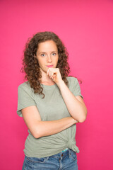 Portrait young redhead girl with hand on chin wearing green t-shirt looking at camera over pink...