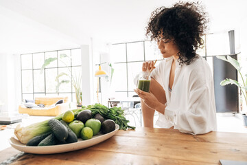 Young african american pretty woman relaxing in kitchen while having a green juice with bamboo...